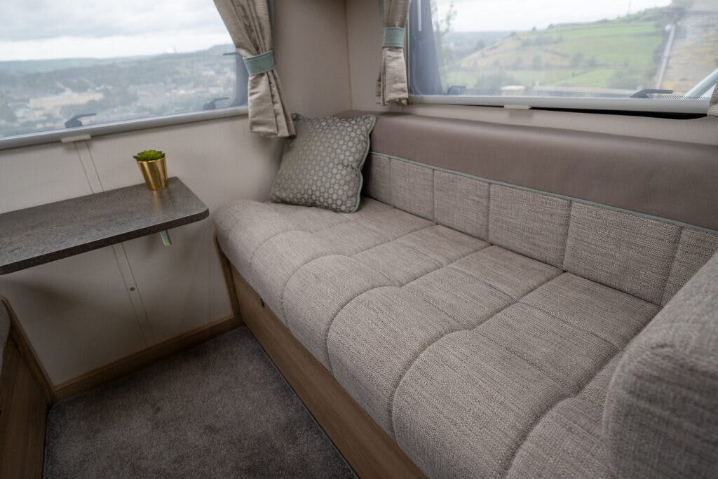 A narrow couch with gray upholstery and a patterned cushion is adjacent to a window in the 2019 Elddis Autoquest 196 Signature Edition RV. A small table with a potted plant sits next to the couch, offering a serene view of the hilly landscape through the window while resting on the carpeted floor.