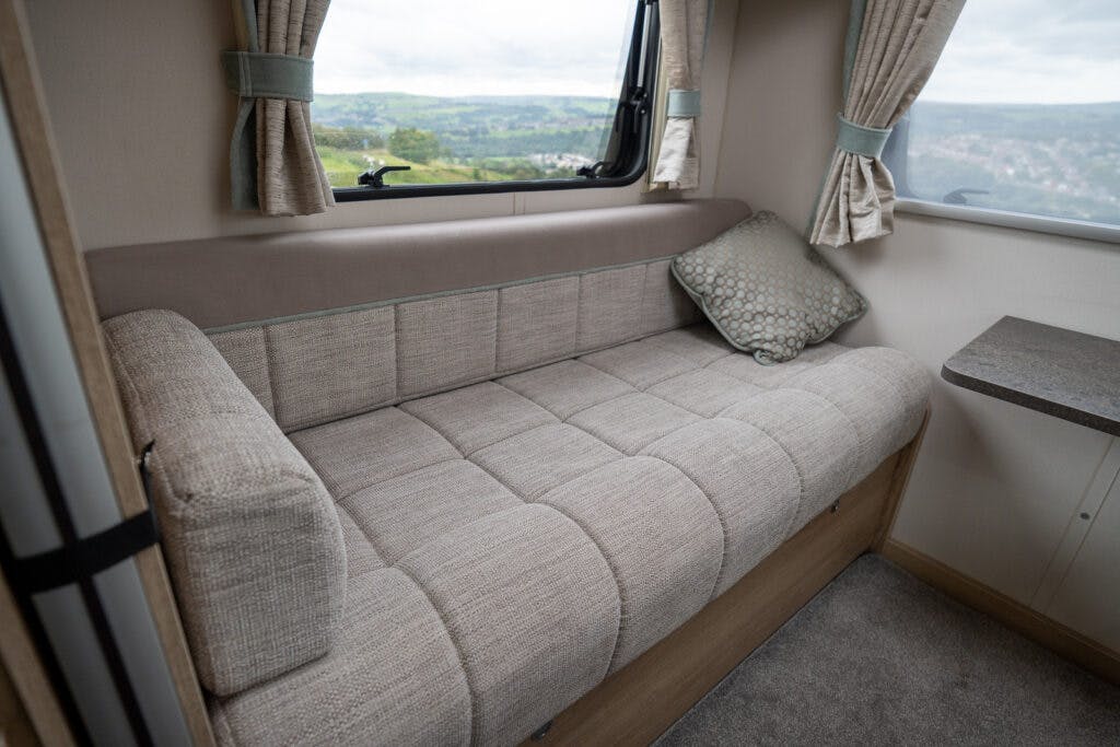 A beige upholstered sofa is situated beneath a window in a well-lit room of the 2019 Elddis Autoquest 196 Signature Edition. The window has beige curtains tied back, offering a view of green hills and a cloudy sky outside. There is a patterned cushion on the sofa and a small countertop beside it.