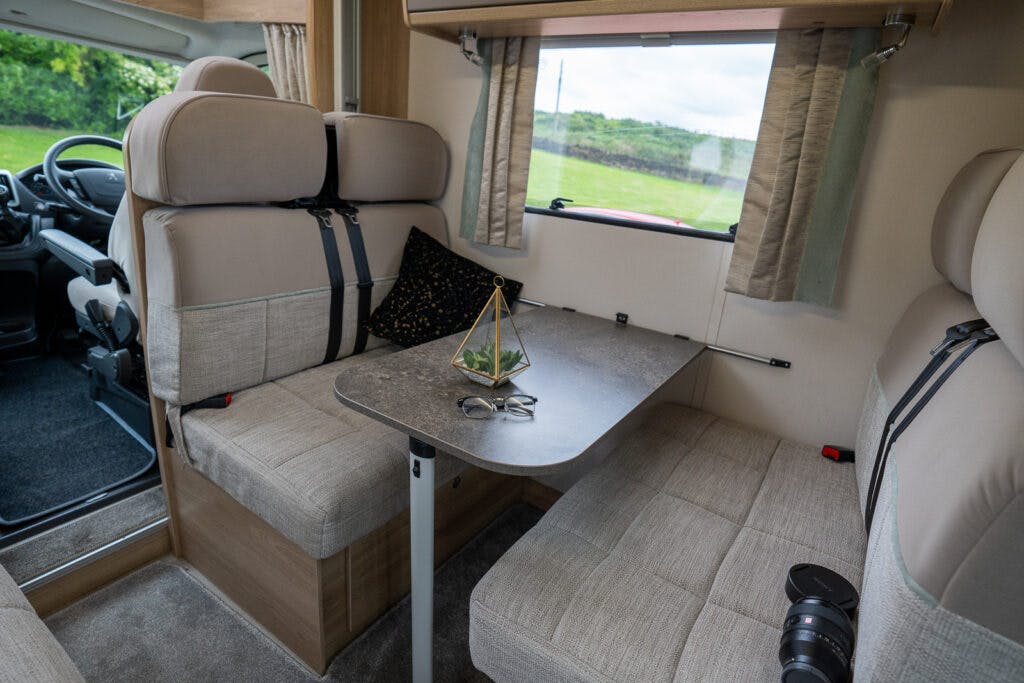 Interior view of the 2019 Elddis Autoquest 196 Signature Edition dining area with beige cushioned seats on either side of a table. On the table, there is a pair of reading glasses, a triangular terrarium, and a camera lens. The RV windows are covered with beige curtains, and the driver's seat is visible.