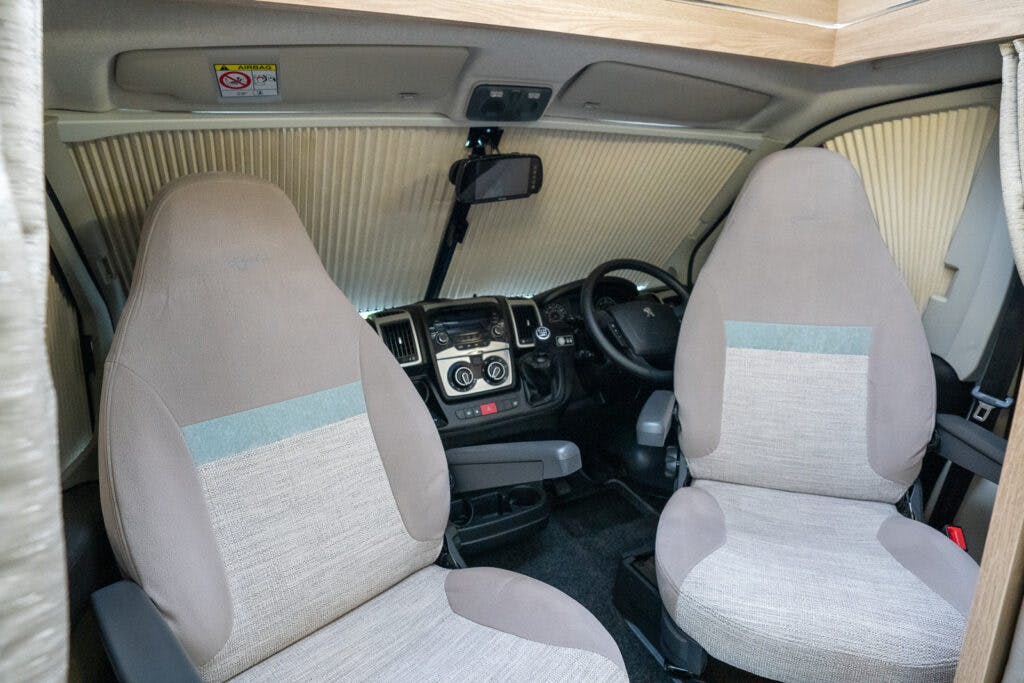 Interior view of the front cabin of the 2019 Elddis Autoquest 196 Signature Edition. The area features two beige upholstered seats with armrests, a black steering wheel, a central dashboard with various controls and a gear stick. Cream-colored pleated blinds are drawn across the windshield.