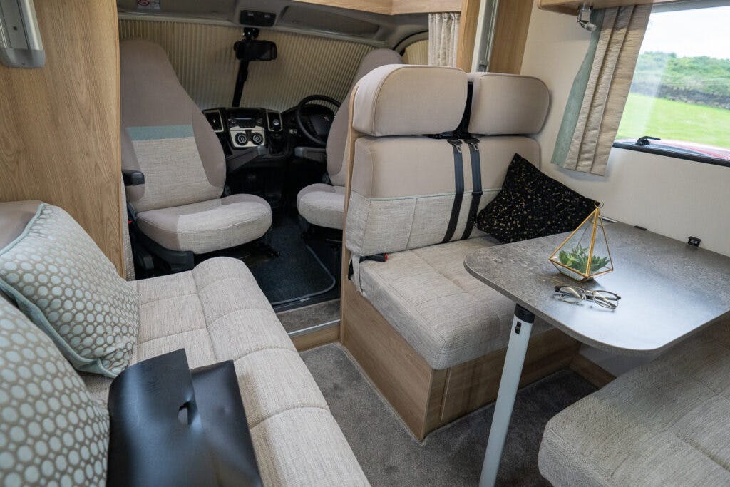 The interior of the 2019 Elddis Autoquest 196 Signature Edition motorhome features a seating area with cream-colored upholstery, a small table adorned with a geometric terrarium and eyeglasses, and the driver's cockpit has two swivel seats. The windows are dressed with curtains, revealing serene green views outside.