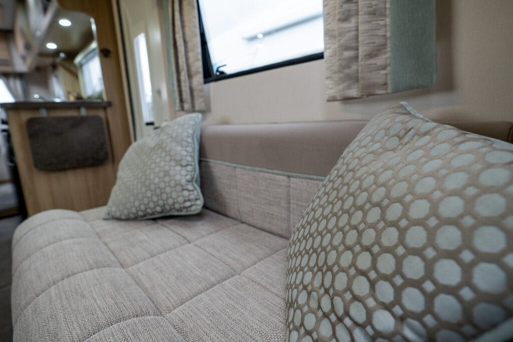 The image shows the interior of what appears to be a 2019 Elddis Autoquest 196 Signature Edition mobile home or RV. It features a beige couch with two decorative pillows in light blue and gray hexagonal patterns. In the background, part of a kitchen area is visible.
