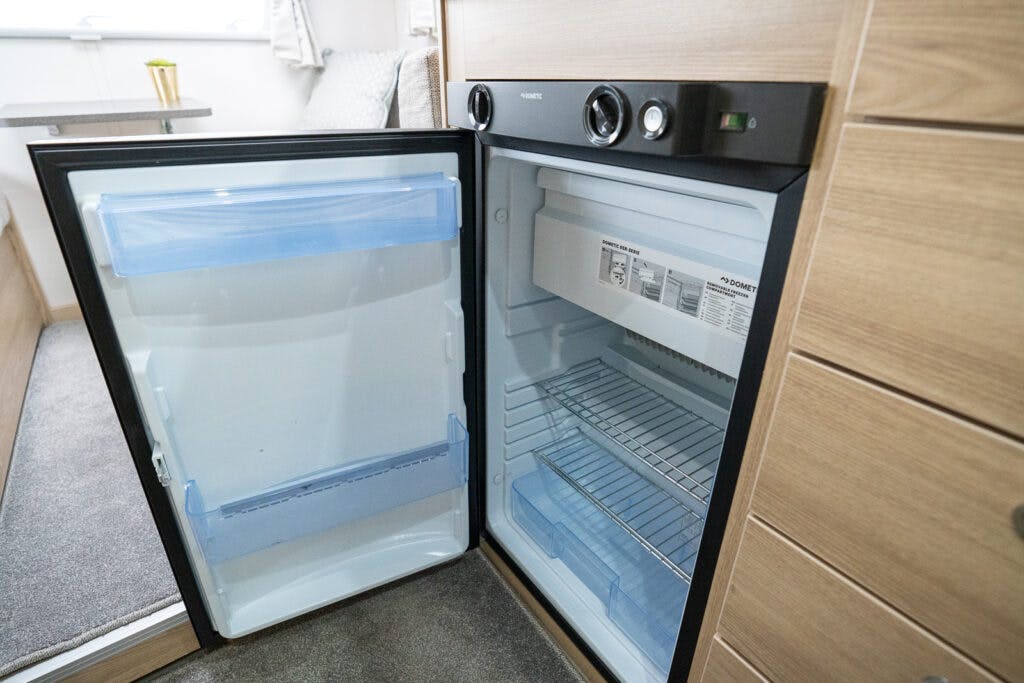 An open mini-fridge with a black frame is shown, reminiscent of the compact appliances found in the 2019 Elddis Autoquest 196 Signature Edition. The inside of the fridge is empty, with three wire shelves and a clear plastic shelf on the door. The control dials and brand information are visible on the upper section of the fridge.