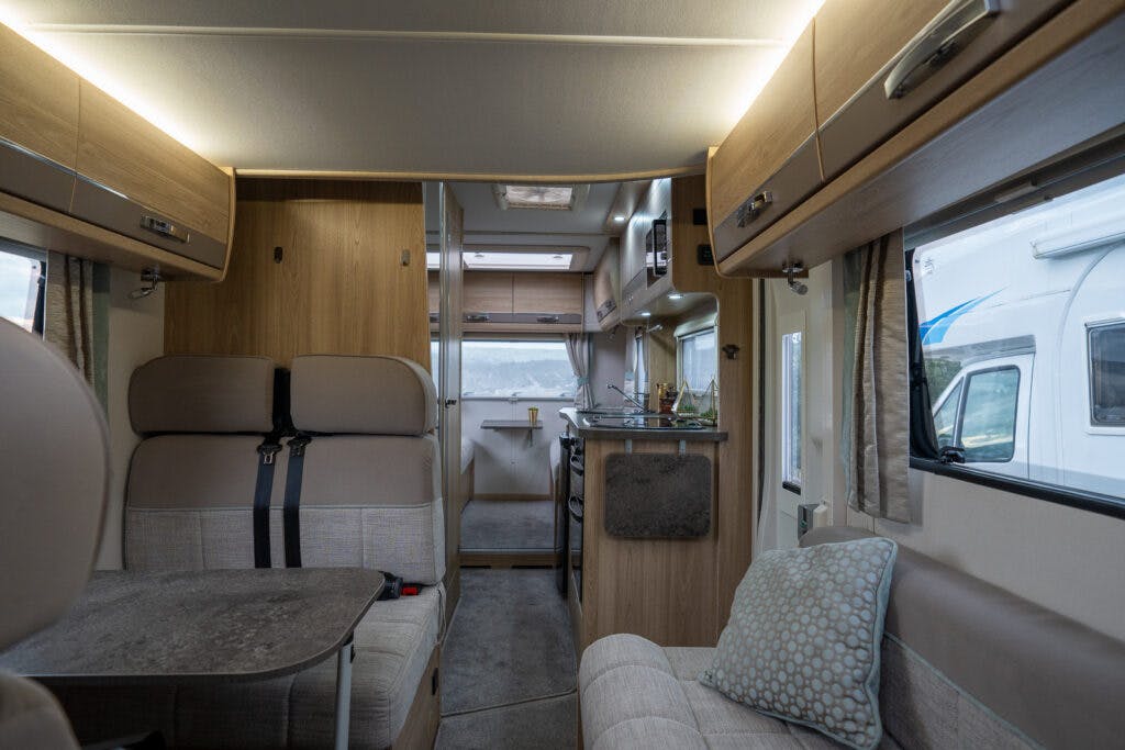 The interior of the 2019 Elddis Autoquest 196 Signature Edition camper van features a seating area with a table on the left and a kitchen on the right. The furniture is wood-toned, with beige upholstery on the seats. The far end of the van has a door and window, letting in natural light.