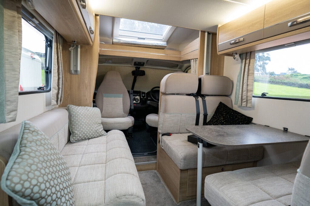 The image shows the interior of a 2019 Elddis Autoquest 196 Signature Edition motorhome. It includes a seating area with beige cushions, a table, and storage cabinets. The driver's and passenger's seats are visible towards the front, with a skylight above. The scene is well-lit and cozy.