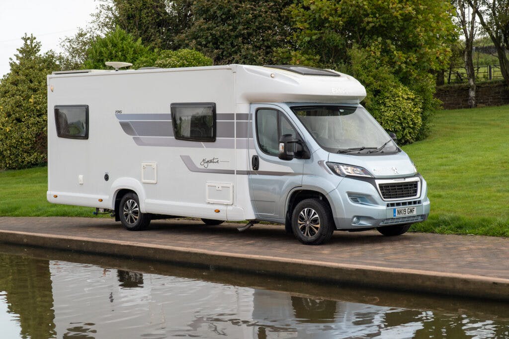 A white and silver 2019 Elddis Autoquest 196 Signature Edition motorhome with the front cab and wheels parked on a brick path next to a body of water. The motorhome features multiple windows, including one with an awning, and green shrubbery and trees are visible in the background.