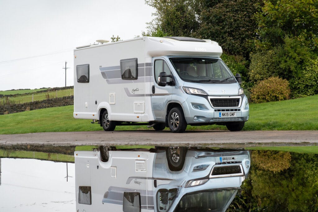 A white and gray 2019 Elddis Autoquest 196 Signature Edition motorhome is parked on a paved surface next to a grassy area with trees. The motorhome's reflection shimmers in a puddle of water in the foreground, and the vehicle's license plate reads "NK51 GHF.