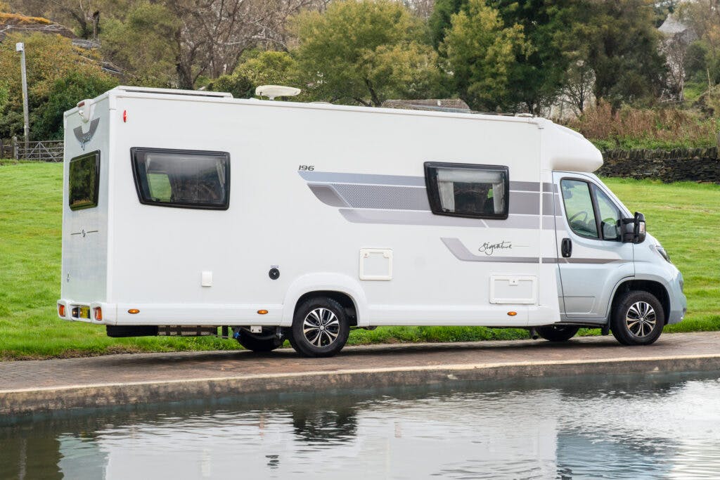 A 2019 Elddis Autoquest 196 Signature Edition motorhome is parked beside a body of water on a paved pathway. The motorhome has dark tinted windows and subtle grey stripes along the side. The background includes grass, trees, and a partially visible fence.