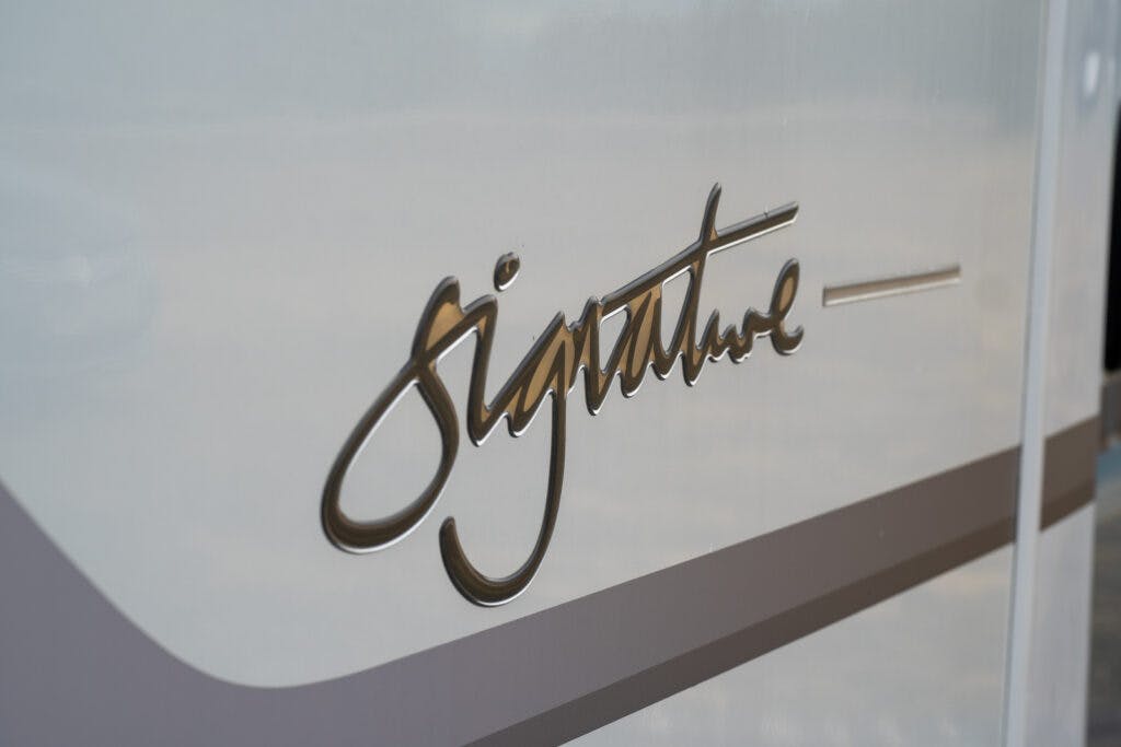 Close-up image of a white surface with the word "Signature" written in cursive golden letters. A thin horizontal stripe also runs parallel to the word, reminiscent of the elegance seen in the 2019 Elddis Autoquest 196 Signature Edition.