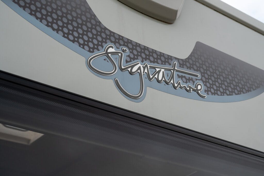 Close-up of a logo on a light-colored, metal surface that reads "Signature" in a stylized, cursive font. The background of the logo features a pattern of small dots arranged in diagonal rows, with parts of the surface in grey and blue hues—reminiscent of the 2019 Elddis Autoquest 196 Signature Edition.