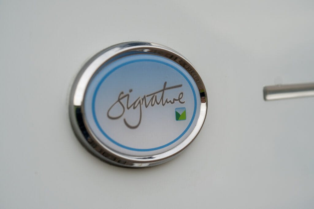 A circular emblem with a blue and white design features the word "signature" in cursive script and a small green and yellow geometric shape in the lower right corner. This distinctive badge, attached to a white surface, marks the 2019 Elddis Autoquest 196 Signature Edition.