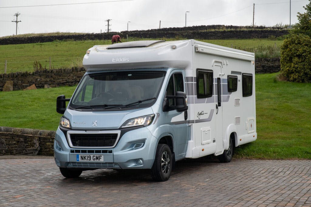 A white and light blue 2019 Elddis Autoquest 196 Signature Edition motorhome is parked on a paved area near a grassy field. It has a UK license plate reading "NK19 GMF" and features various windows and storage compartments. Power lines and a stone fence can be seen in the background.