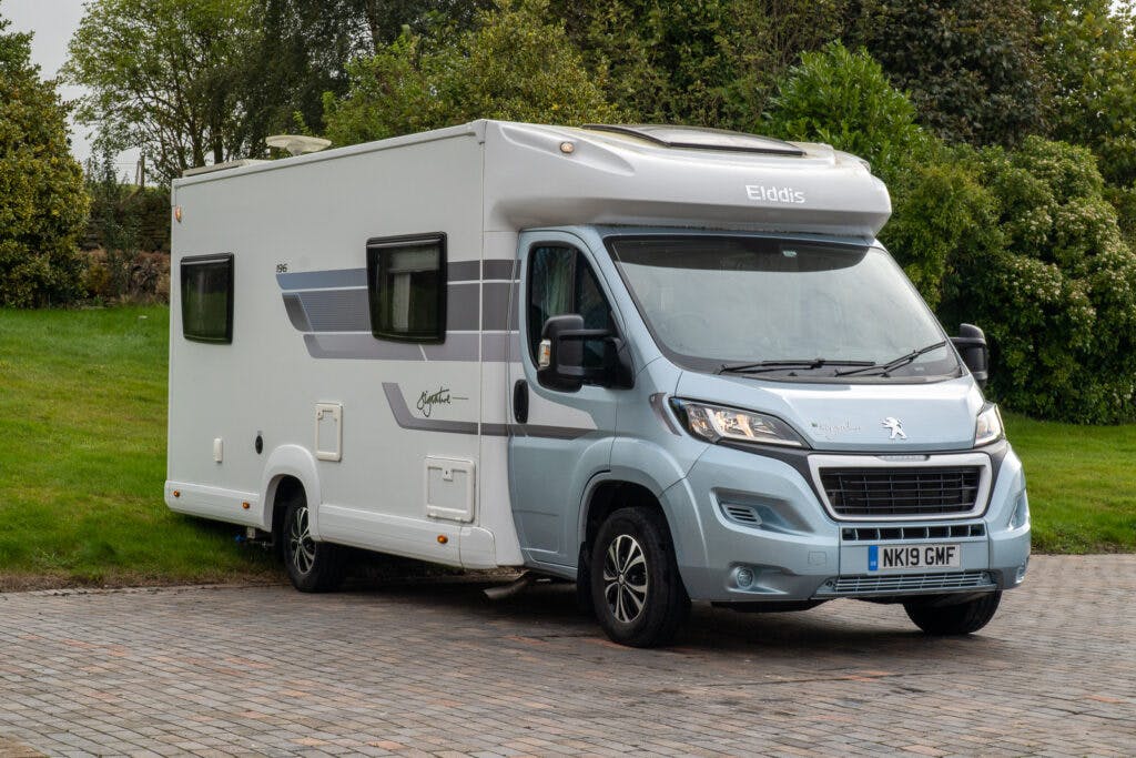 A 2019 Elddis Autoquest 196 Signature Edition motorhome is parked on a paved area with green grass and trees in the background. The vehicle, with UK license plate number NK19 GMF, is white and light blue, featuring several windows and a Peugeot chassis.