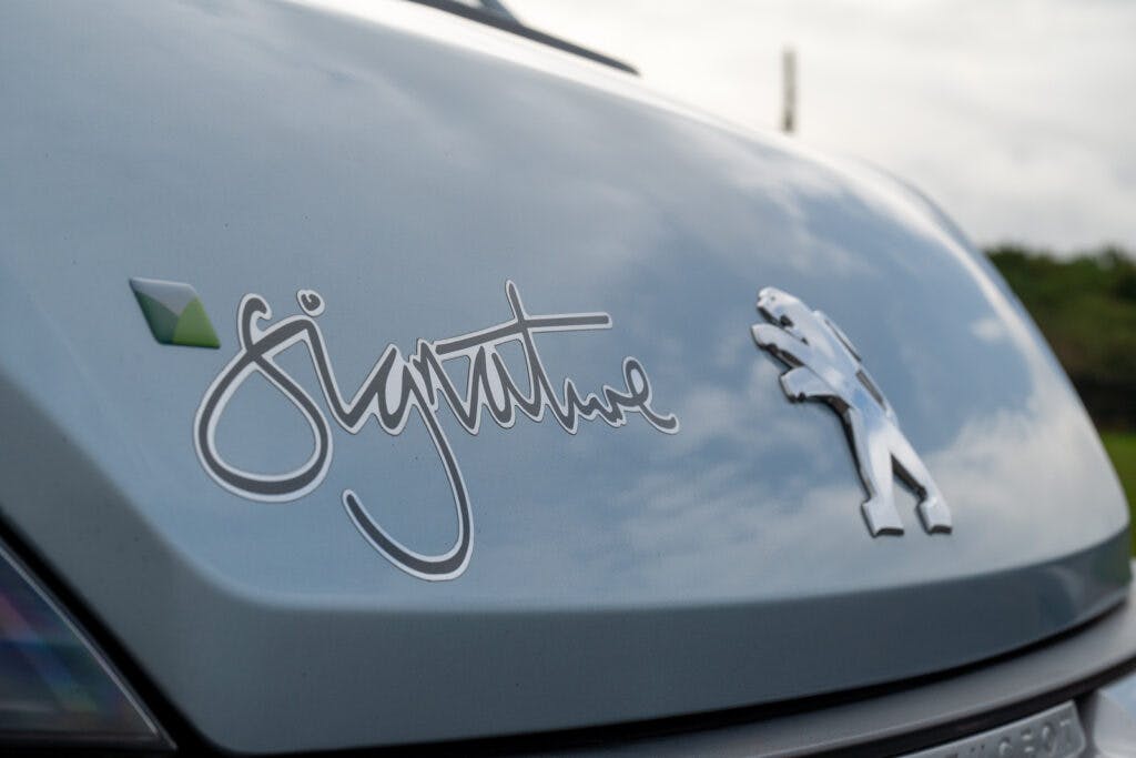 Close-up image of a car's hood featuring the Peugeot logo and the word "Signature" in cursive text. The car, which is part of the 2019 Elddis Autoquest 196 Signature Edition, appears to be blue, and the background shows a cloudy sky and some greenery.