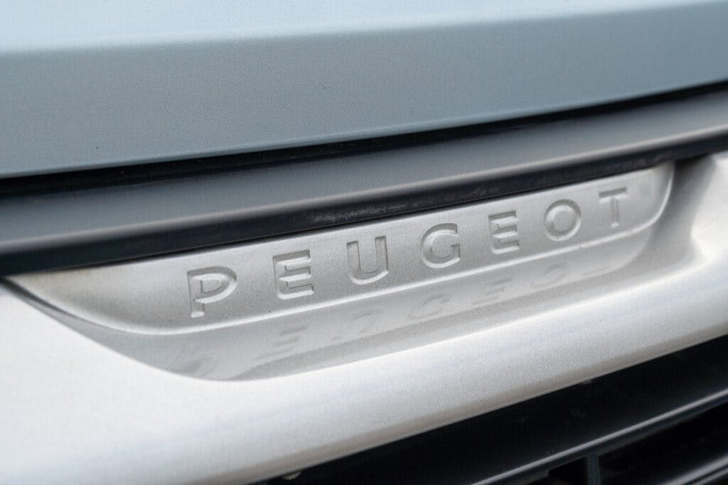 Close-up of a Peugeot car logo on the front grille, displaying the brand name embossed in metallic letters against a silver background. The image focuses on the central part of the grille with part of the 2019 Elddis Autoquest 196 Signature Edition's front end visible.