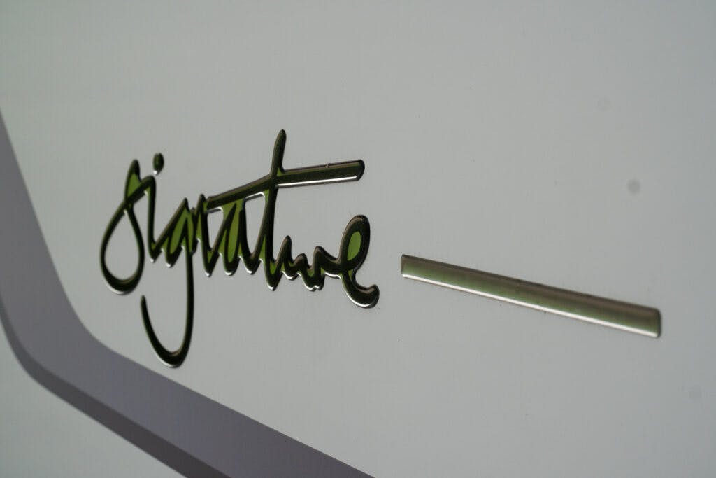 A close-up image of the gold-colored word "signature" written in cursive on a smooth, light gray surface. A horizontal gold line extends from the right end of the word, reminiscent of the elegant branding found on the 2019 Elddis Autoquest 196 Signature Edition.