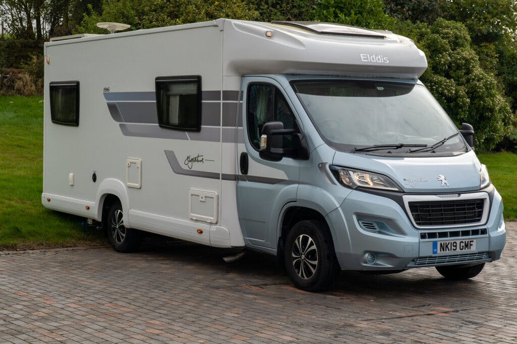 A white and blue 2019 Elddis Autoquest 196 Signature Edition motorhome is parked on a paved area. The vehicle, with a registered number plate "NK19 GMF," features tinted windows, side mirrors, and various branding elements on its exterior. Greenery is visible in the background.