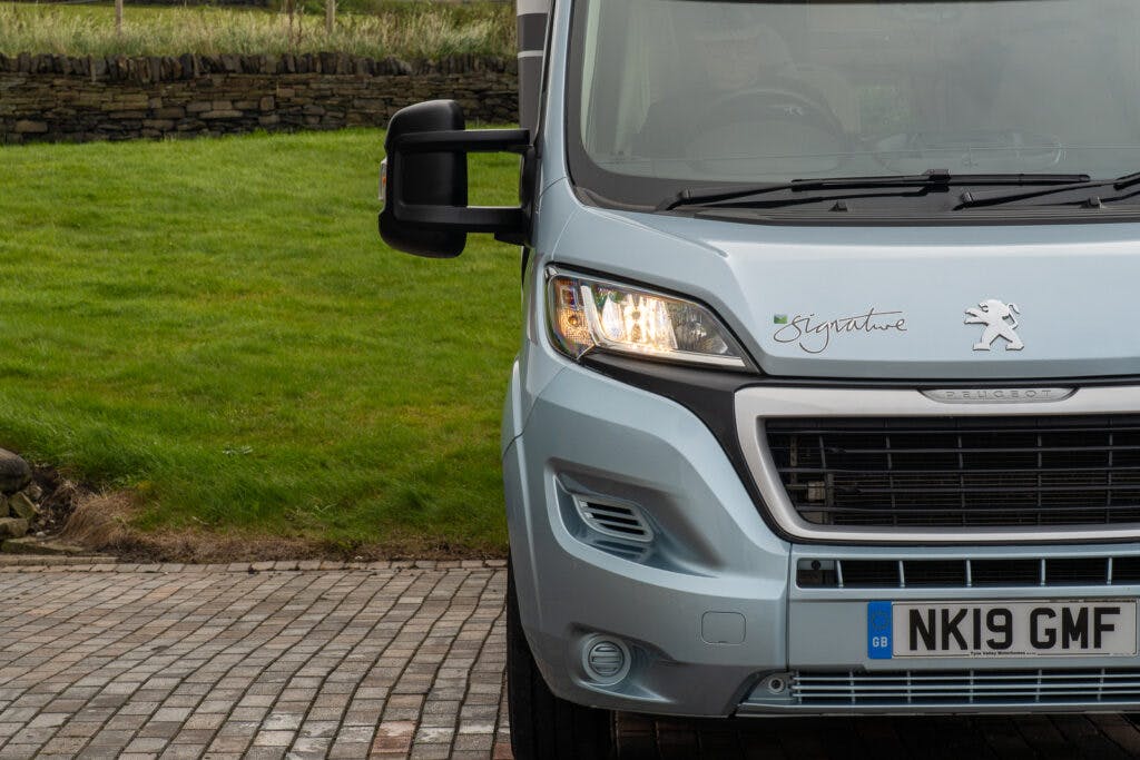 A front view of a Peugeot vehicle with a lit headlight and a license plate reading "NK19 GMF." The vehicle, part of the 2019 Elddis Autoquest 196 Signature Edition, is parked on a paved area with green grass and a stone wall in the background.