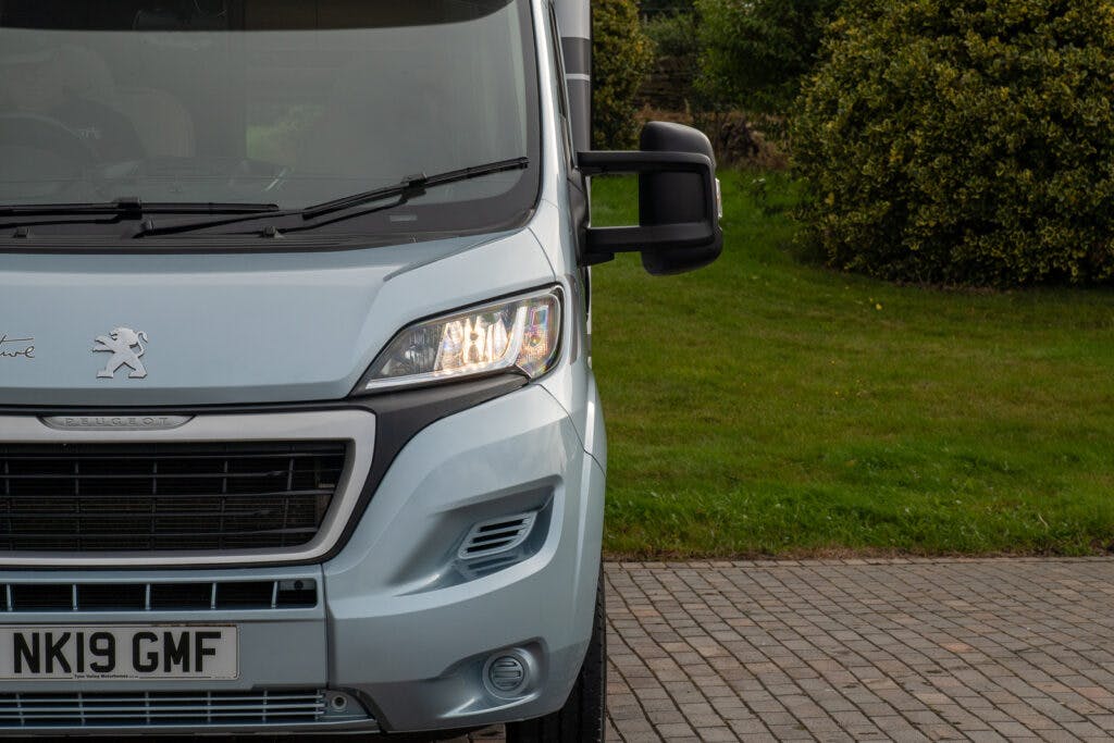 The front left side of a silver Peugeot van, possibly a 2019 Elddis Autoquest 196 Signature Edition, is shown parked on a driveway. The van's left headlight is illuminated, and the side view mirror is visible. The license plate reads "NK19 GMF." A grassy area and trees are in the background.