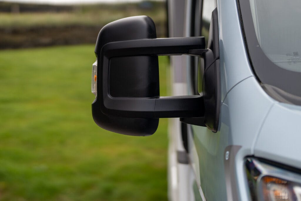 Close-up of a side mirror on a 2019 Elddis Autoquest 196 Signature Edition with a grassy field in the background. The mirror is large and rectangular with a black casing, complementing the light blue vehicle.