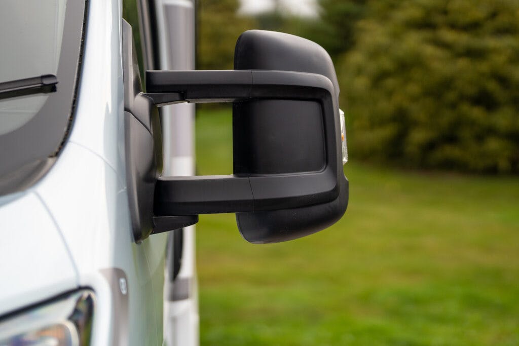 A close-up side view of a black side mirror on a white 2019 Elddis Autoquest 196 Signature Edition. The background shows green grass and blurred bushes.
