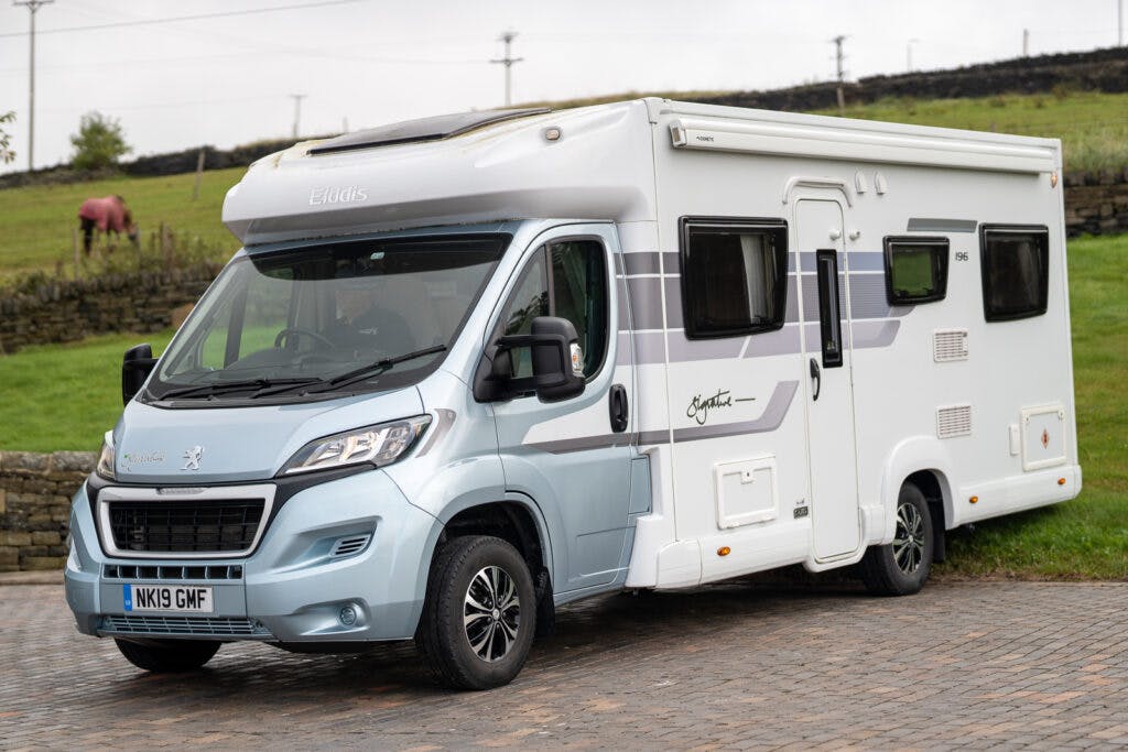 A white and light blue 2019 Elddis Autoquest 196 Signature Edition motorhome is parked on a paved area. The vehicle has a Peugeot base, and the license plate reads "NK19 GMF." In the background, a grassy field with stone walls and a grazing horse can be seen.