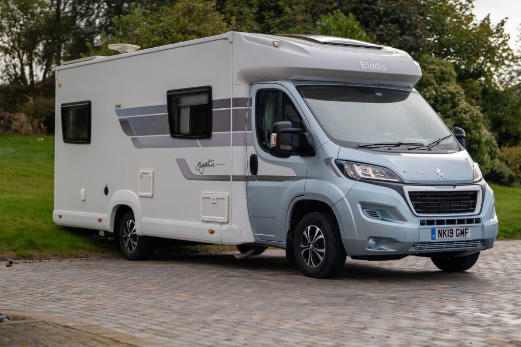 This image shows a parked 2019 Elddis Autoquest 196 Signature Edition motorhome. The vehicle features a white and grey exterior with a Peugeot front cab. It is situated on a paved area with green grass and trees visible in the background. The license plate reads "NK19 GMF.