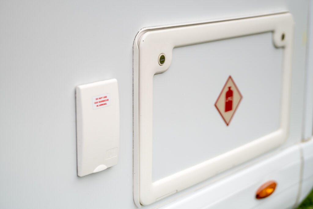 Close-up of a 2019 Elddis Autoquest 196 Signature Edition's exterior side panel, featuring a rectangular storage compartment with a small red gas cylinder warning symbol. A smaller, white rectangular component is positioned to the left of the compartment, featuring a red text label.
