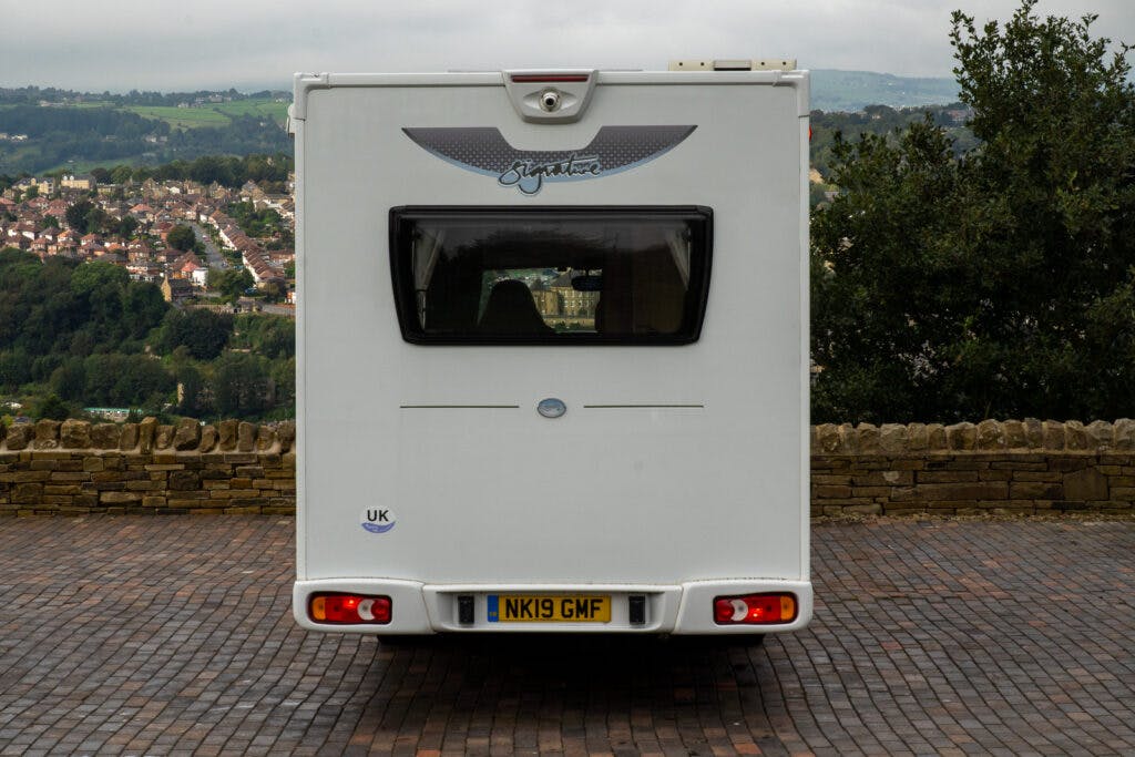 A white 2019 Elddis Autoquest 196 Signature Edition camper van is parked on a cobblestone surface with its rear facing the camera. The vehicle, bearing a UK license plate "NK19 GMF," features a "Signature" logo. In the background, there's a scenic view of a hilly town with houses and greenery.