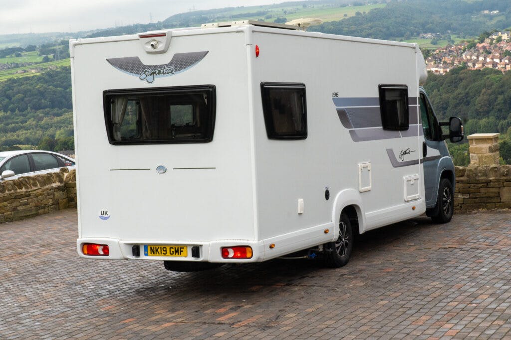 A 2019 Elddis Autoquest 196 Signature Edition motorhome is parked on a stone-paved area overlooking a valley with trees and distant buildings. The vehicle, with multiple windows, a UK sticker, and a license plate reading "NK19 GMF," stands under an overcast sky.