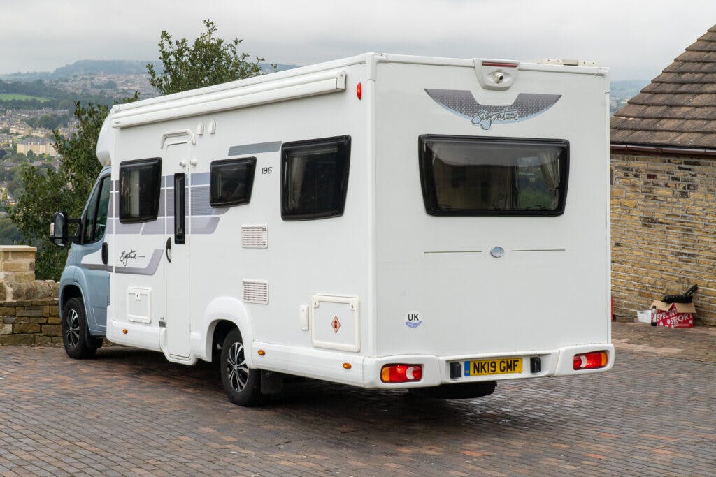 A white 2019 Elddis Autoquest 196 Signature Edition motorhome, displaying registration plate NK19 GMF, is parked on a brick driveway. The back of the vehicle shows various windows, a rear camera, and branding. A stone wall, trees, and a house with a tiled roof are in the background.