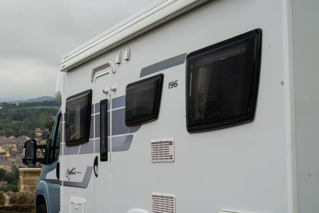 A close-up view of the side of a 2019 Elddis Autoquest 196 Signature Edition RV, labeled with the number 196. The image shows two black-tinted windows and a partially visible landscape with hills and buildings in the background on an overcast day.