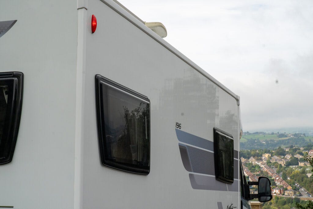 A partial side view of the 2019 Elddis Autoquest 196 Signature Edition RV with dark narrow windows. The exterior is white with grey and blue graphics, and a small red light is visible on the upper left. The background reveals a distant landscape with houses and greenery.