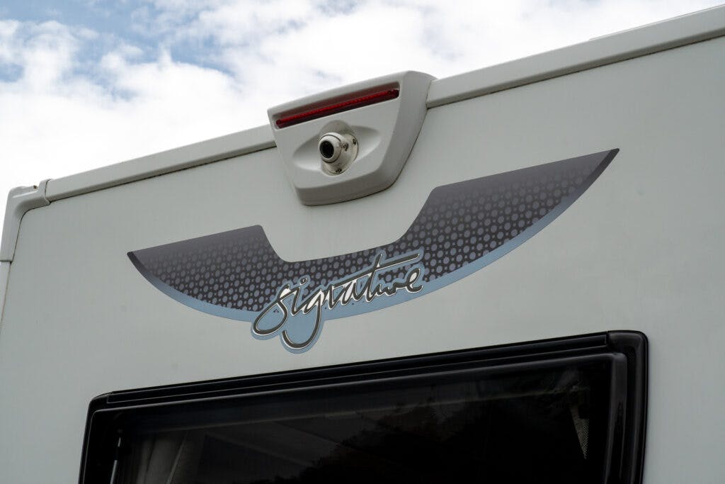 Close-up of the rear section of a white 2019 Elddis Autoquest 196 Signature Edition camper van. The image shows a decorative decal with the word "Signature" and a white rear camera mounted above it. There is a window below the decal, and the sky in the background is partly cloudy.