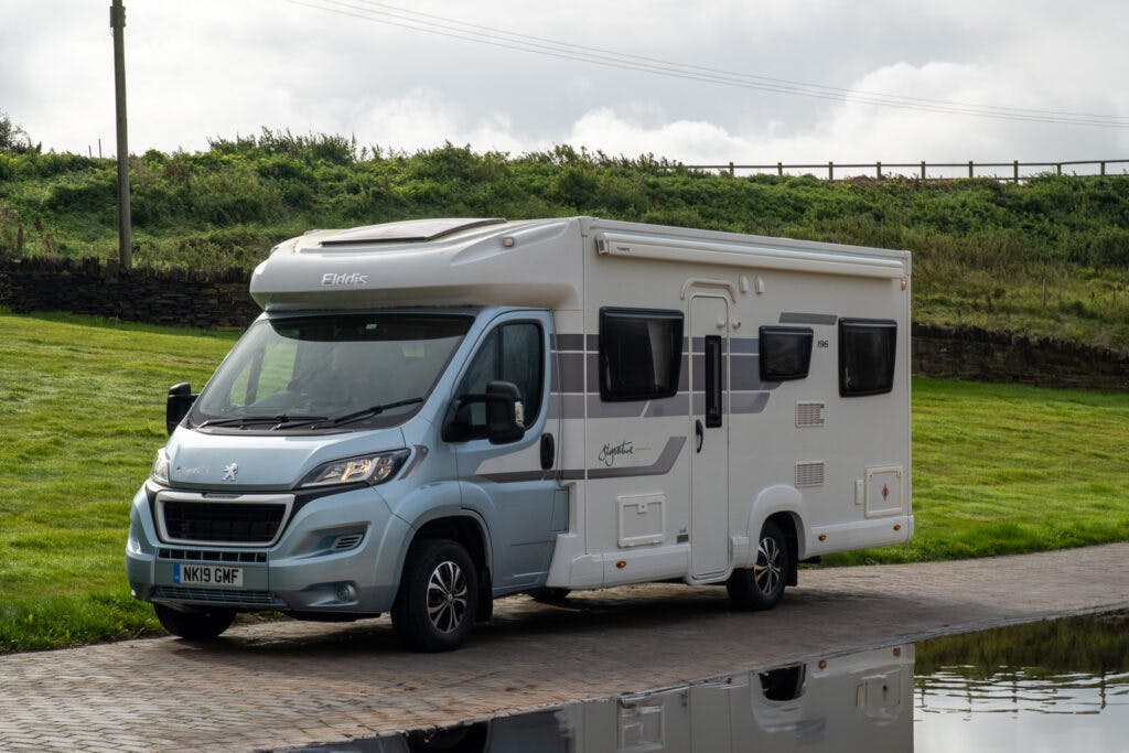 A gray and white 2019 Elddis Autoquest 196 Signature Edition motorhome is parked on a paved area next to a canal. The vehicle features large windows and a Peugeot logo on the front. In the background is a grassy hill with a wooden fence running along its top, under a partly cloudy sky.