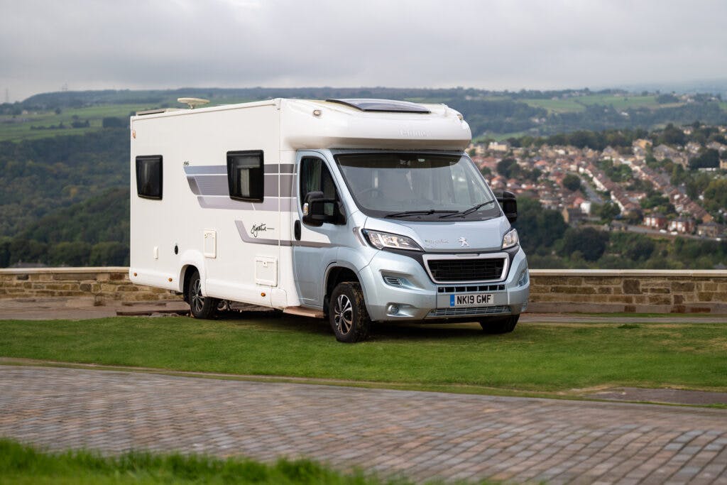 A white 2019 Elddis Autoquest 196 Signature Edition motorhome with license plate number NK19 GWF is parked on a grassy area. Behind it is a stone railing and a view of a town with houses, greenery, and hills under a cloudy sky.