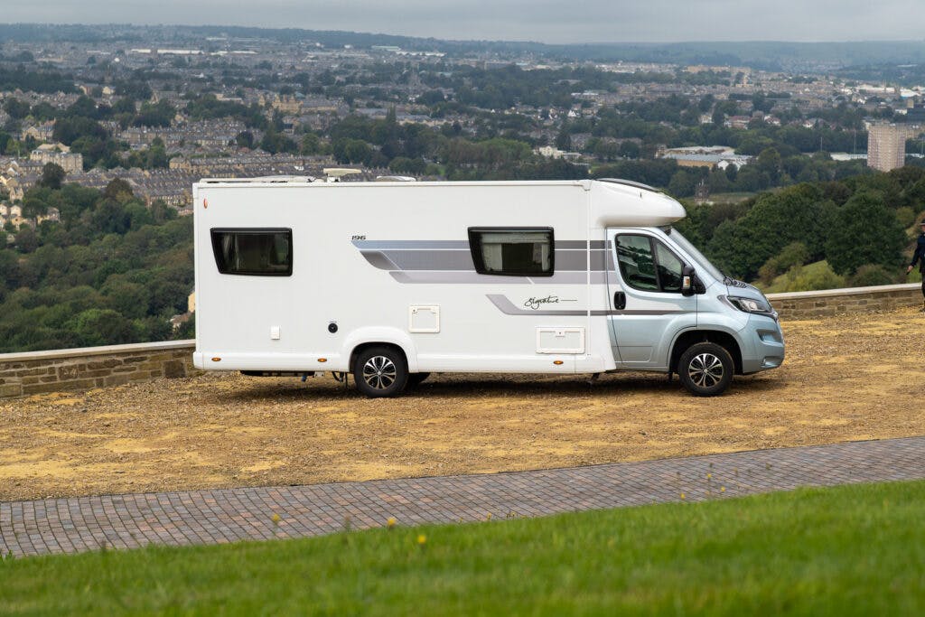 A white 2019 Elddis Autoquest 196 Signature Edition camper van is parked on a gravel area with a city skyline and green hills in the background. The sky is overcast, and the van has various windows and markings on its side.