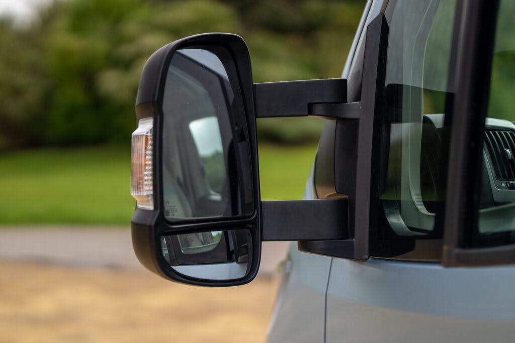 Close-up of a side mirror on the 2019 Elddis Autoquest 196 Signature Edition, showcasing both the main mirror and the smaller convex mirror below it. The background includes blurred green foliage and a hint of pavement.