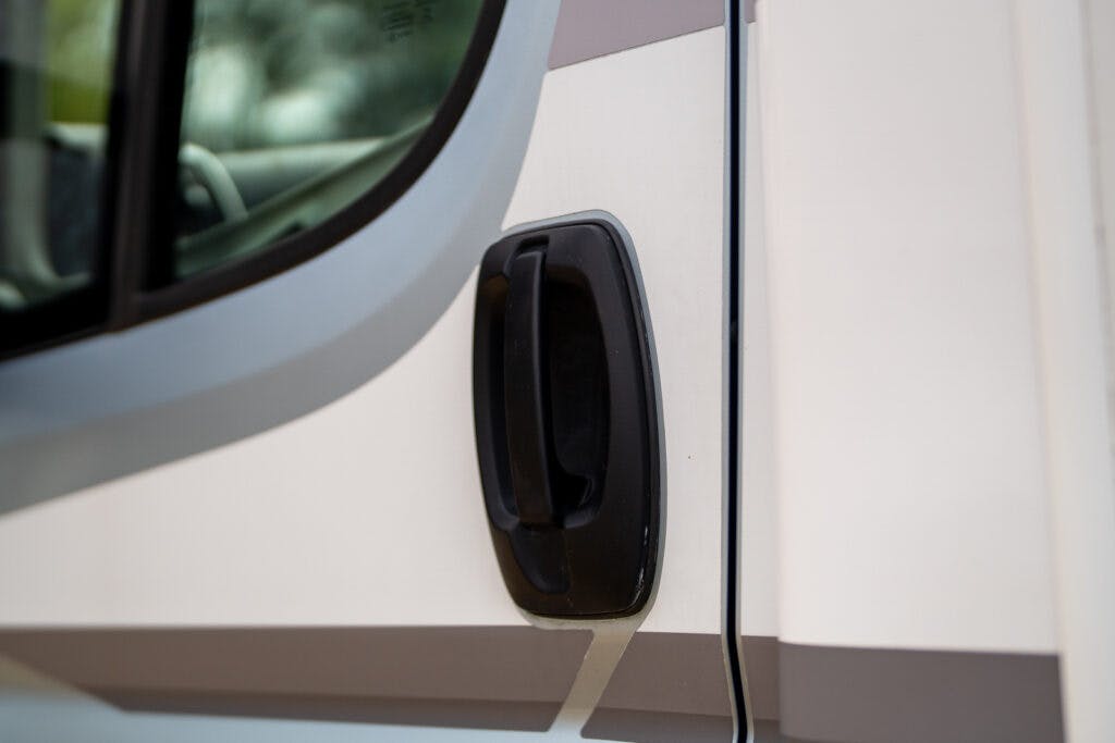 Close-up of a black handle on the exterior of a white vehicle, likely a van or truck. The handle is positioned near a window with slightly tinted glass, capturing part of the 2019 Elddis Autoquest 196 Signature Edition’s door.