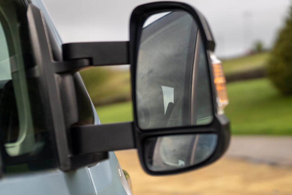 Close-up view of a 2019 Elddis Autoquest 196 Signature Edition’s side mirror showing the side and rear view. The mirror, mounted on the driver's side, reflects part of the surrounding landscape with greenery and a cloudy sky. There's a slight reflection of the vehicle's interior.