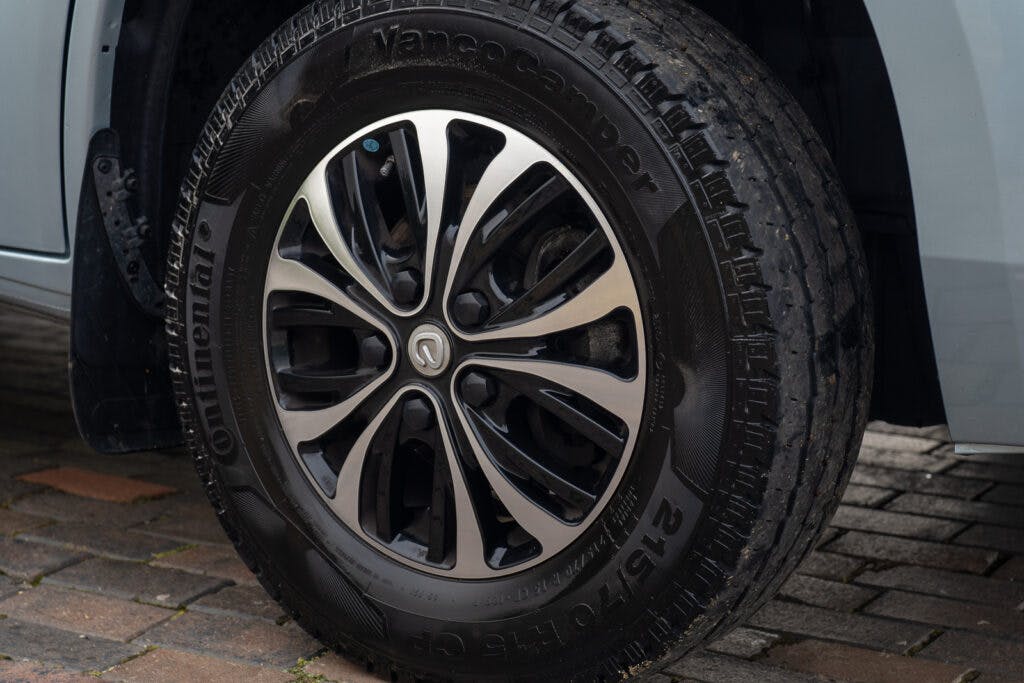 Close-up of a car tire and wheel on a stone-paved surface. The tire is a Continental VanContact, ideal for the 2019 Elddis Autoquest 196 Signature Edition, with an all-season tread pattern. The wheel boasts a modern, multi-spoke design with a polished metallic finish.