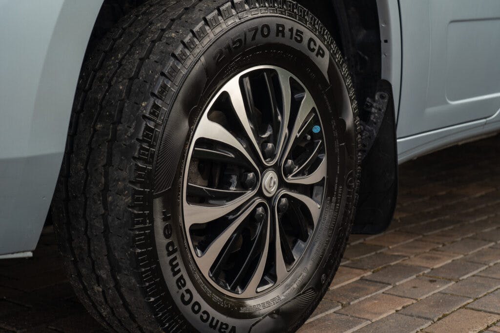 A close-up of the front tire on the 2019 Elddis Autoquest 196 Signature Edition, showcasing the detailed tread pattern and modern alloy wheel design. The tire has the specifications "215/70 R15 CP" and the brand "VancoCamper" imprinted on its sidewall. The car is parked on a brick surface.