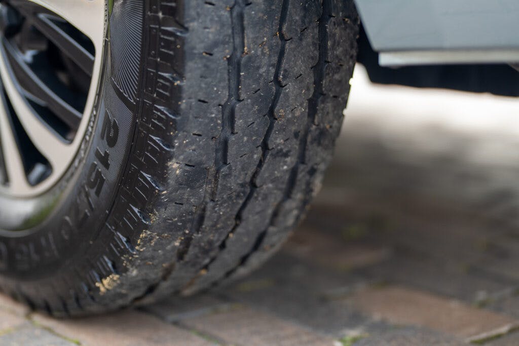 Close-up of a worn car tire with visible tread wear, indicating the need for replacement. The tire is shown on a brick-paved surface beside the 2019 Elddis Autoquest 196 Signature Edition, with the bottom part of the vehicle visible in the background.