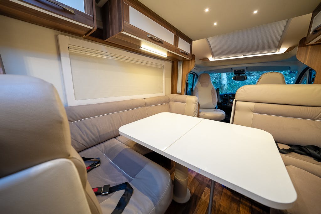 Interior of the 2016 Roller Team Auto-Roller 707 Low Line motorhome with beige and brown tones. The space includes cushioned seating around a white table, seatbelts on the seats, cabinetry above, and a view of the driver's area in the distance.