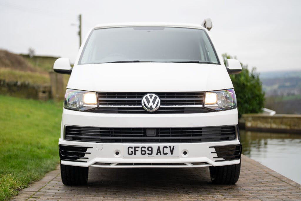 A white 2019 Volkswagen Transporter T28 Trendline TDI van with its headlights on is parked facing forward on a paved path. The vehicle, featuring a license plate reading "GF69 ACV," is surrounded by greenery and set against a cloudy sky backdrop.