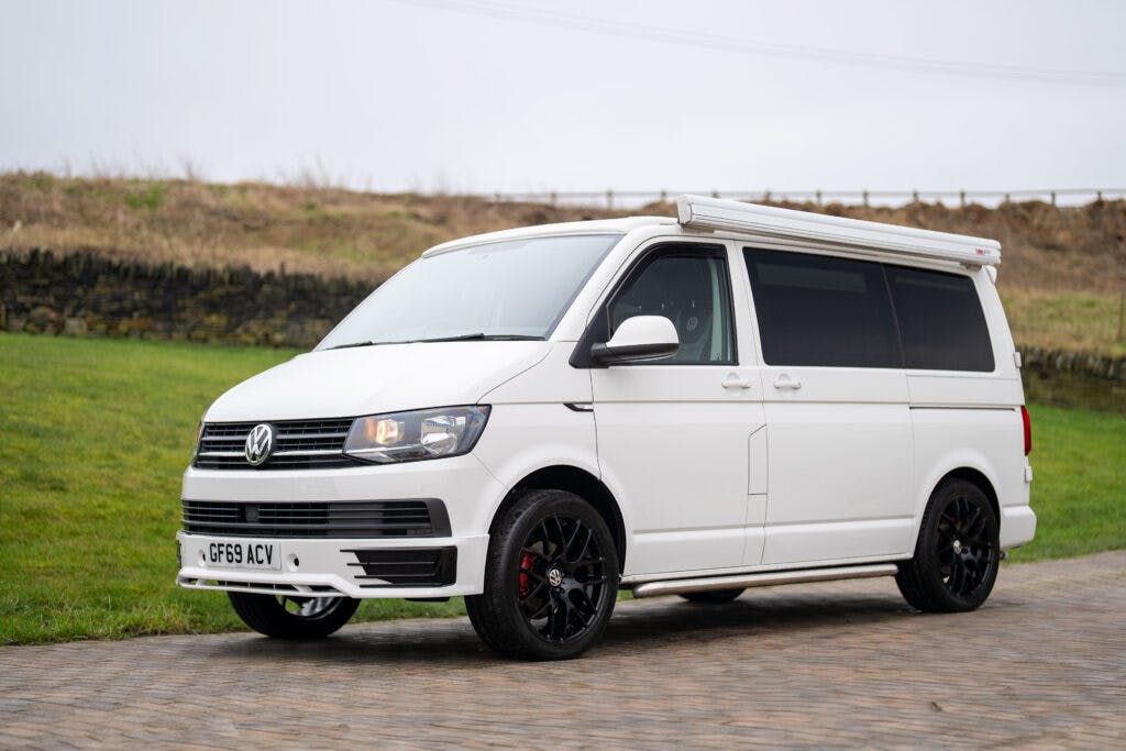 A white 2019 Volkswagen Transporter T28 Trendline TDI is parked on a paved road with a grassy hill and stone wall in the background. The van features black wheels, tinted windows, and a retractable roof canopy. The license plate reads GF69 ACV.