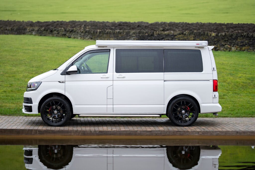A 2019 Volkswagen Transporter T28 Trendline TDI is parked on a paved path in front of a grassy field and a stone wall. The van, with its roof box and black wheels, casts a crisp reflection in the body of water in the foreground.