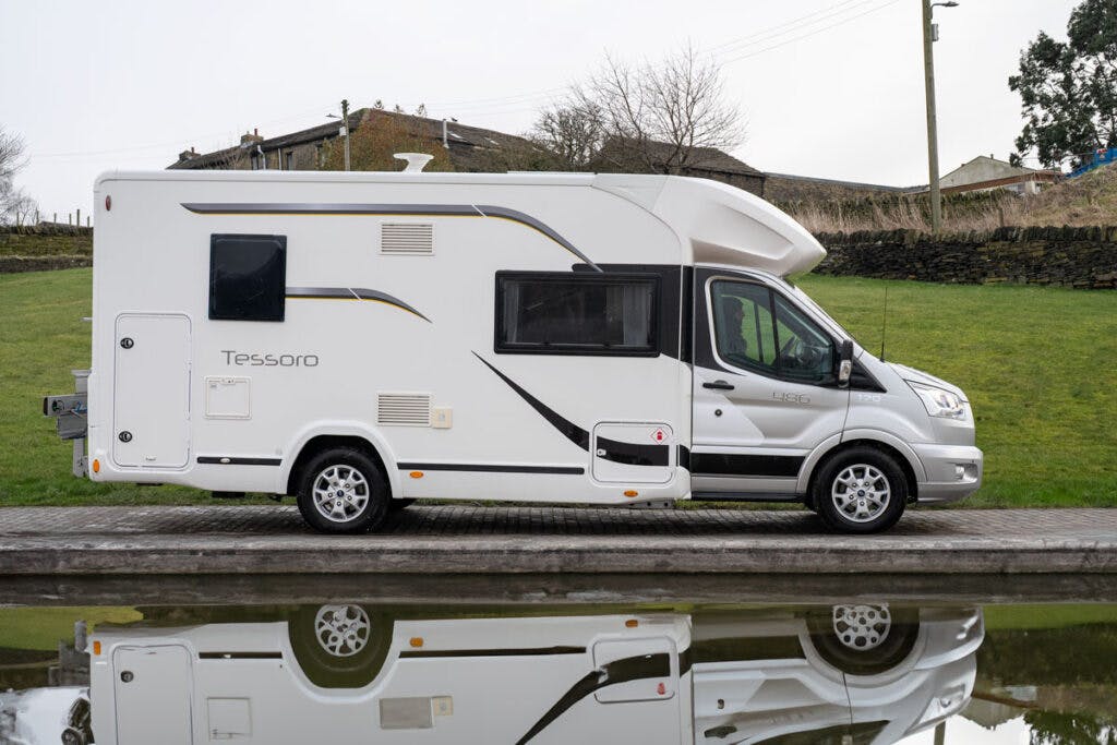 A 2019 Benimar Tessoro T486 motorhome is parked on a paved surface beside a calm body of water, reflecting its image. The surroundings include grass, a stone wall, and some houses on a slightly elevated area in the background.