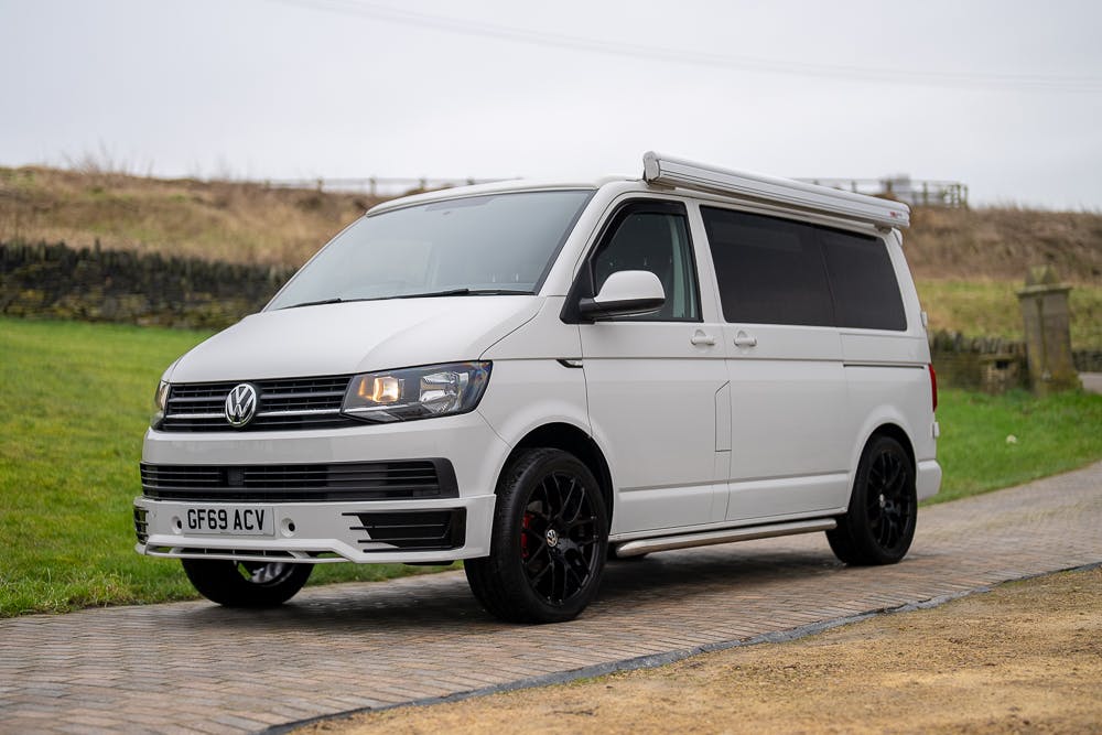 A white 2019 Volkswagen Transporter T28 Trendline TDI camper van is parked on a paved and grassy area. The van has black wheels, tinted windows, and a license plate that reads "GF69 ACV." The area is open with some fencing and an overcast sky.
