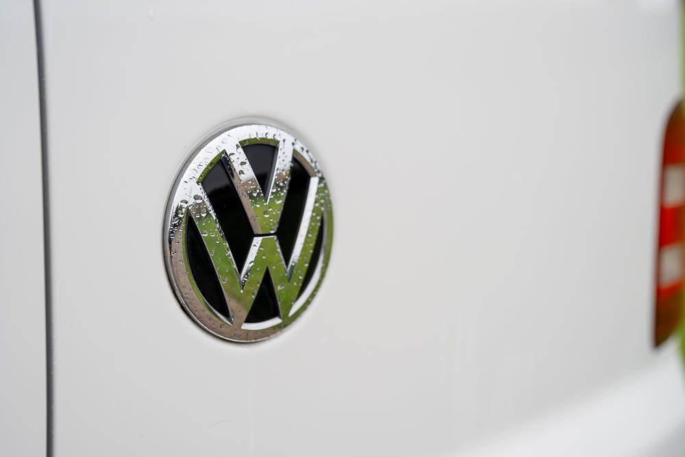 Close-up of the Volkswagen logo on a white 2019 Volkswagen Transporter T28 Trendline TDI. The circular emblem features interlocked silver letters V and W, with water droplets visible on its surface.